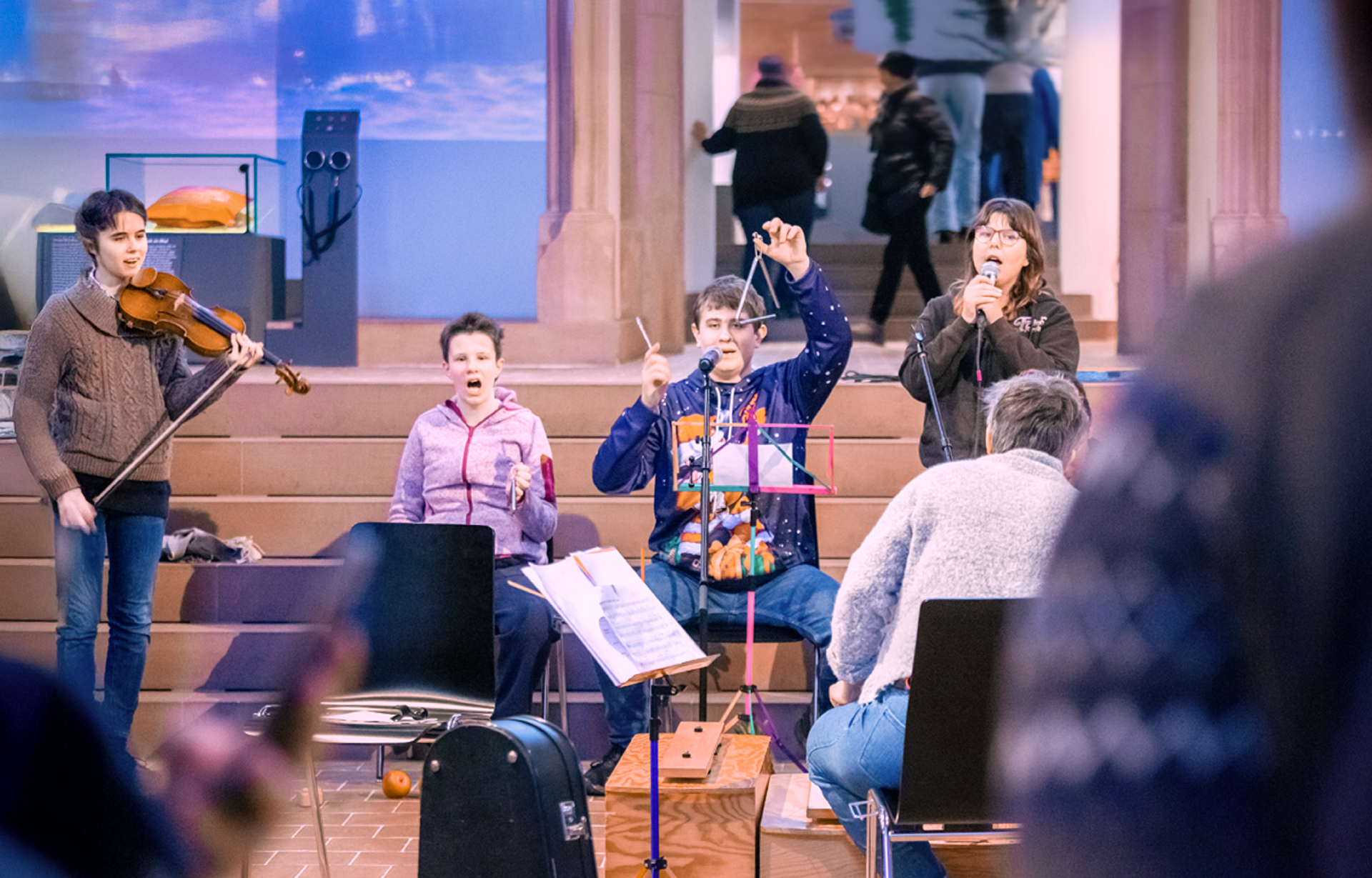 Vier Musiker treten drinnen auf: Einer spielt Violine, zwei singen, und einer spricht in ein Mikrofon. Notenblätter und Instrumente sind sichtbar, mit einem Zuschauer im Vordergrund.